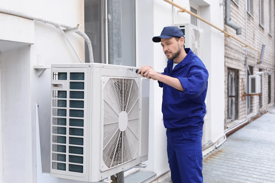 Technicien en uniforme bleu et casquette effectuant la maintenance d'une unité extérieure de climatisation sur terrasse d'immeuble résidentiel